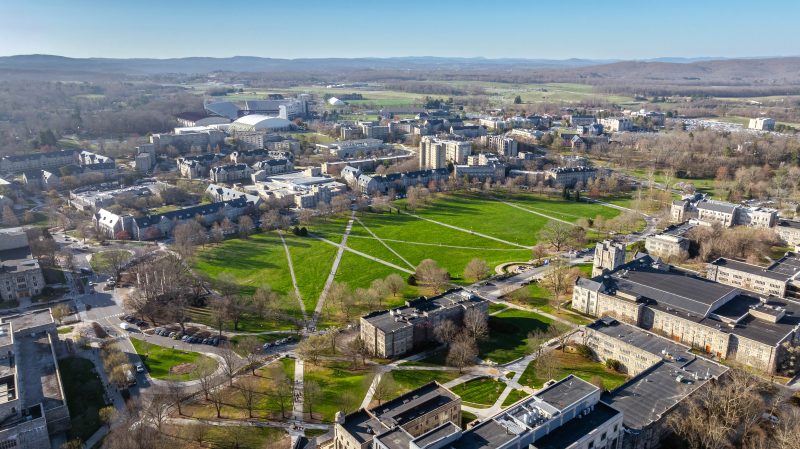 Aerial view of Virginia Tech's Blacksburg campus via drone in the springtime featuring the Drillfield surrounded by budding trees and many grey Hokie Stone buildings