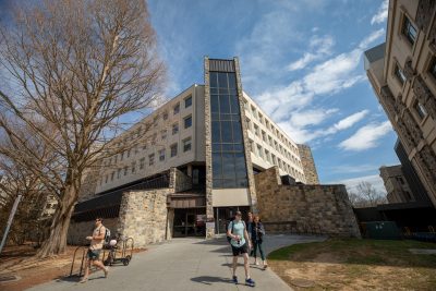 Grey Hokie Stone and concrete McBryde Hall on a sunny spring day. Several people are exiting the building and a large tree with no leaves on it sits to the building's left.